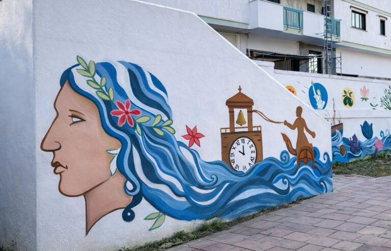 Murale su muro esterno con profilo femminile dai capelli blu trasformati in onde, elementi floreali e simboli legati al mare e al territorio.