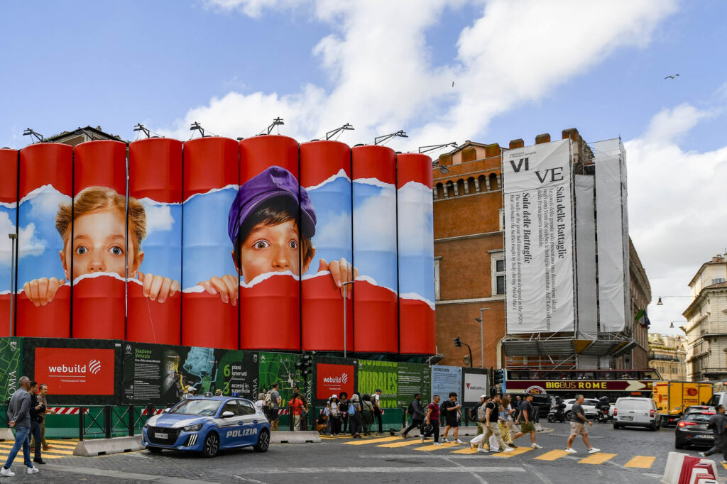 Murale con i volti di due bambini che osservano oltre una superficie rossa, realizzato sulla recinzione del cantiere della stazione Venezia della Metro C a Roma.
