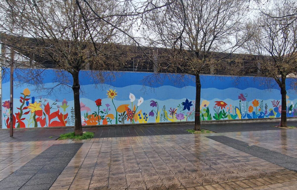 Murale floreale realizzato su una lunga parete urbana, con grandi fiori colorati su fondo azzurro e persone al lavoro durante l’intervento artistico.