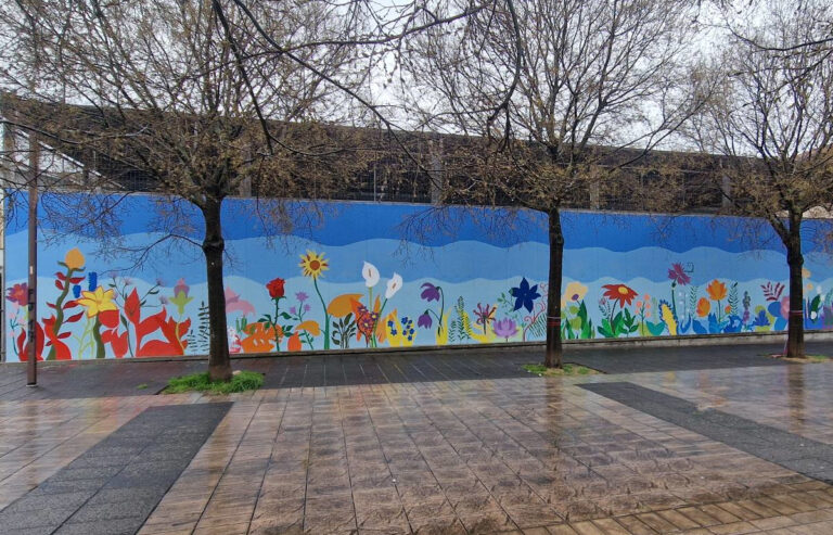 Murale floreale realizzato su una lunga parete urbana, con grandi fiori colorati su fondo azzurro e persone al lavoro durante l’intervento artistico.