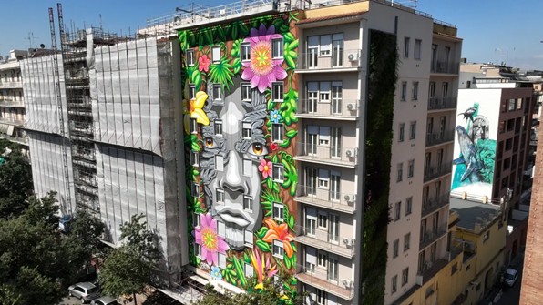 Veduta complessiva del murale Eco Ivy realizzato sulla facciata di un edificio di otto piani a Roma, con un volto monumentale circondato da fiori e foglie tropicali.