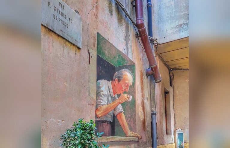 Murale di Eduardo Relero a Fiuggi raffigurante un anziano con i capelli grigi e una camicia chiara che si affaccia da una finestra dipinta, sorseggiando una bevanda da una piccola tazzina bianca. L'opera crea un effetto tridimensionale sulla parete di un vicolo del centro storico.